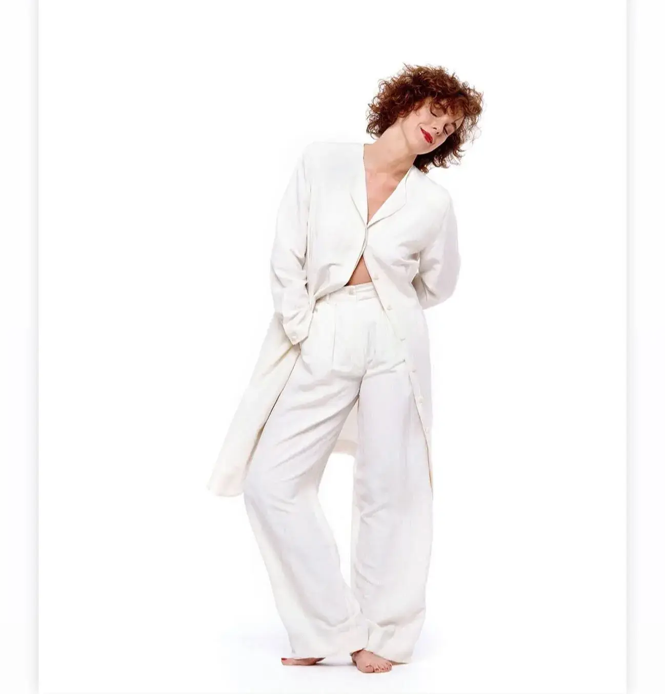 American actress Elizabeth Perkins poses barefoot in a relaxed white suit against a plain white background, tilting her head with her eyes closed in a serene expression.