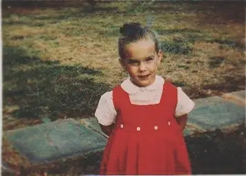 A childhood photo of actress Elizabeth Perkins shows her as a young girl outdoors wearing a red jumper over a white collared shirt.