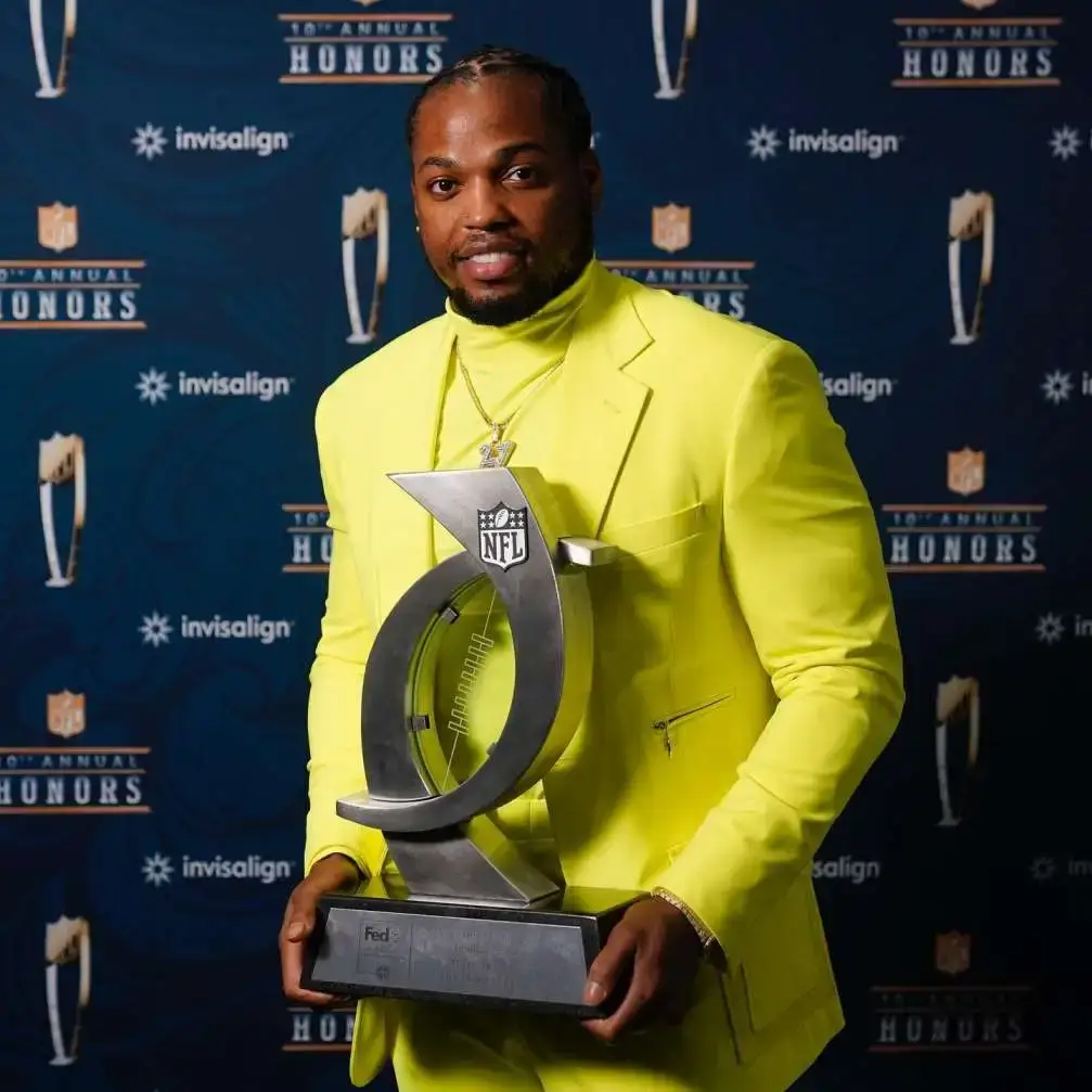 NFL running back Derrick Henry stands in front of a blue "10th Annual Honors" backdrop while wearing a bright neon yellow suit and holding a large silver trophy.