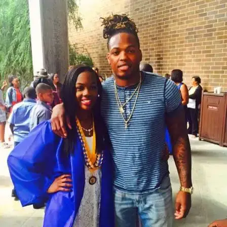 NFL player Derrick Henry stands with his arm around a young woman in a blue graduation gown and yellow honors stole in front of a brick building.