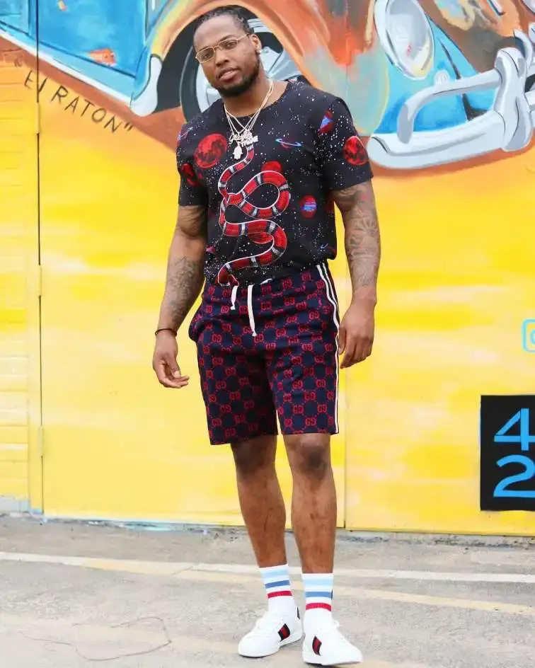 NFL Derrick Henry stands in front of a colorful mural wearing a graphic snake-print t-shirt, patterned shorts, and white sneakers.