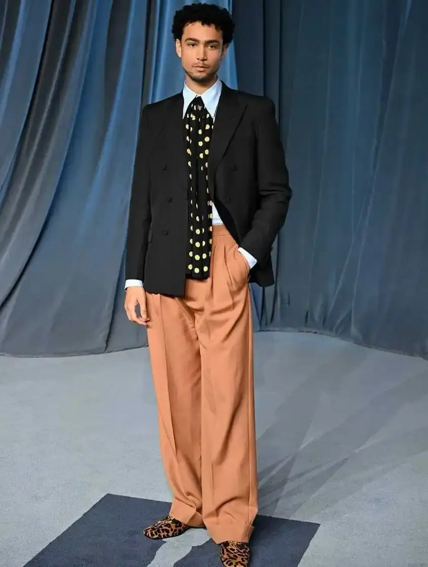 Actor Archie Madekwe poses in a black double-breasted blazer, tan wide-leg trousers, a polka-dot tie, and leopard-print shoes against a draped blue backdrop.