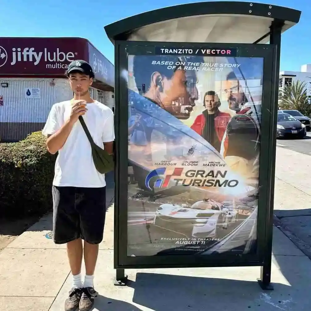 Actor Archie Madekwe stands on a sunny sidewalk wearing a white t-shirt, black shorts, and a baseball cap while drinking from a cup next to a large bus shelter advertisement for his film Gran Turismo.