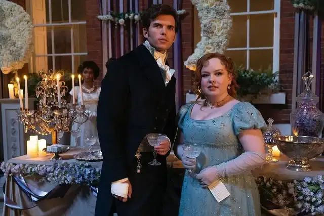 Luke Newton and Nicola Coughlan stand together in Regency-era attire as Colin Bridgerton and Penelope Featherington during a candlelit ballroom scene.