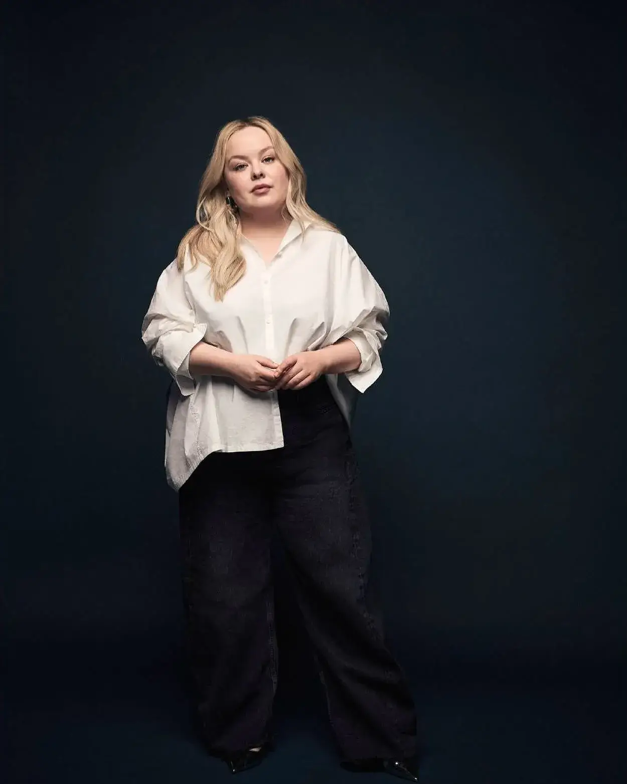Nicola Coughlan poses against a dark blue backdrop wearing an oversized white button-down shirt paired with wide-leg dark denim trousers.