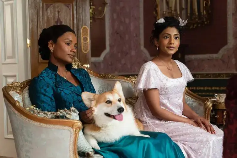 Simone Ashley and Charithra Chandran sit on an ornate sofa in Regency-era gowns with a Corgi during a scene from Bridgerton.