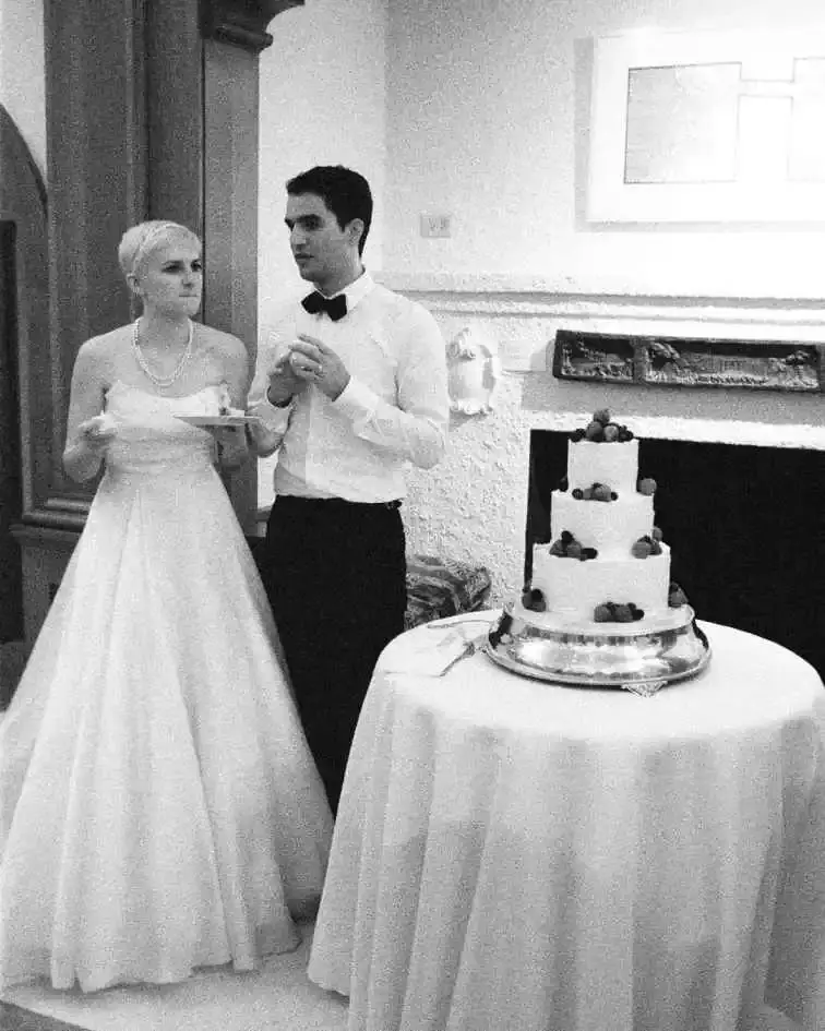 A black and white photo of Benny Safdie in a tuxedo and his wife Ava Safdie in a strapless wedding gown standing beside their three-tiered wedding cake.