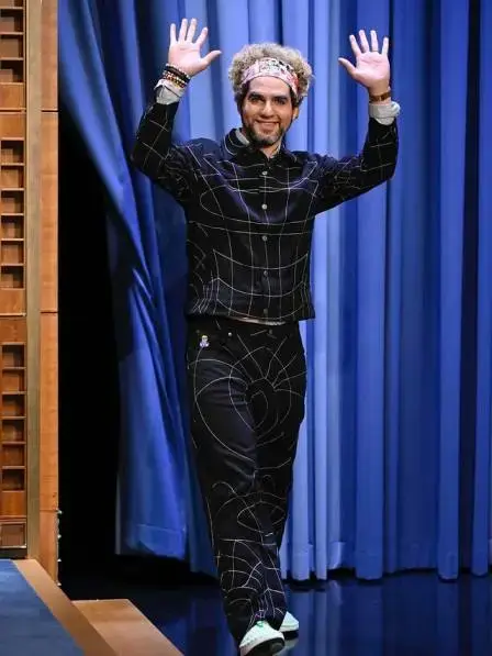 Benny Safdie waves to an audience while wearing a black button-down shirt and matching trousers with a white geometric line pattern.