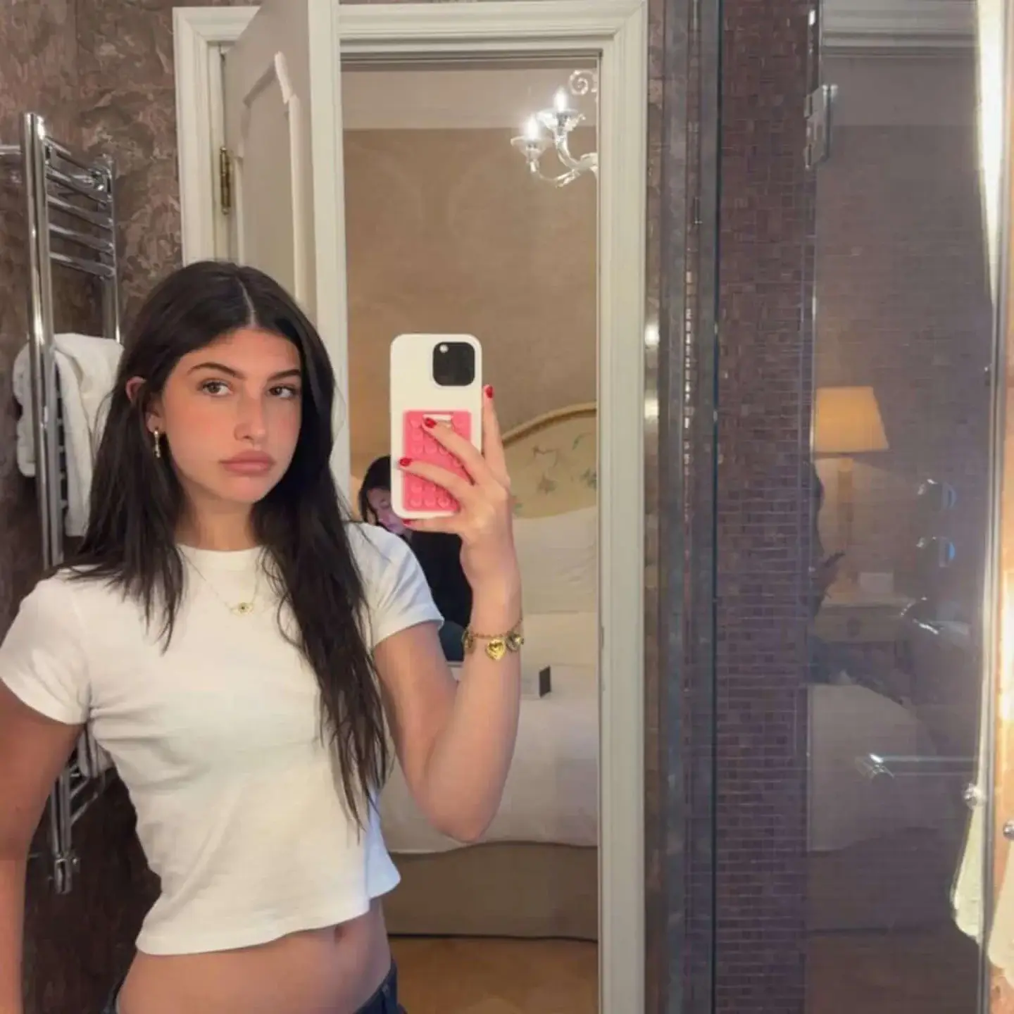 Sadie Sandler takes a mirror selfie in a bathroom wearing a white cropped t-shirt and a gold charm bracelet.