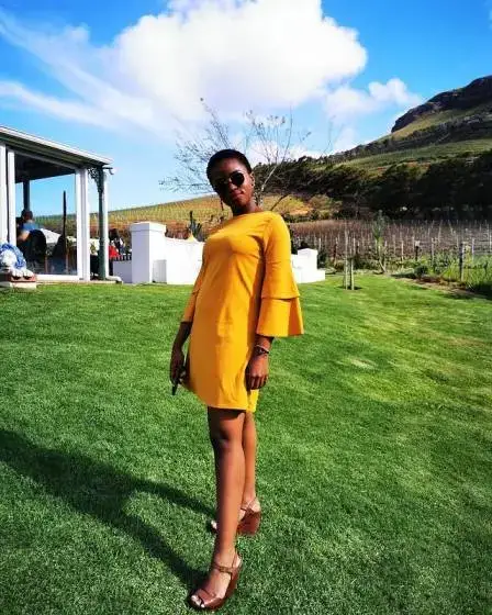 Masali Baduza wearing a yellow bell-sleeve dress and sunglasses while standing on a grassy field with hills in the background.