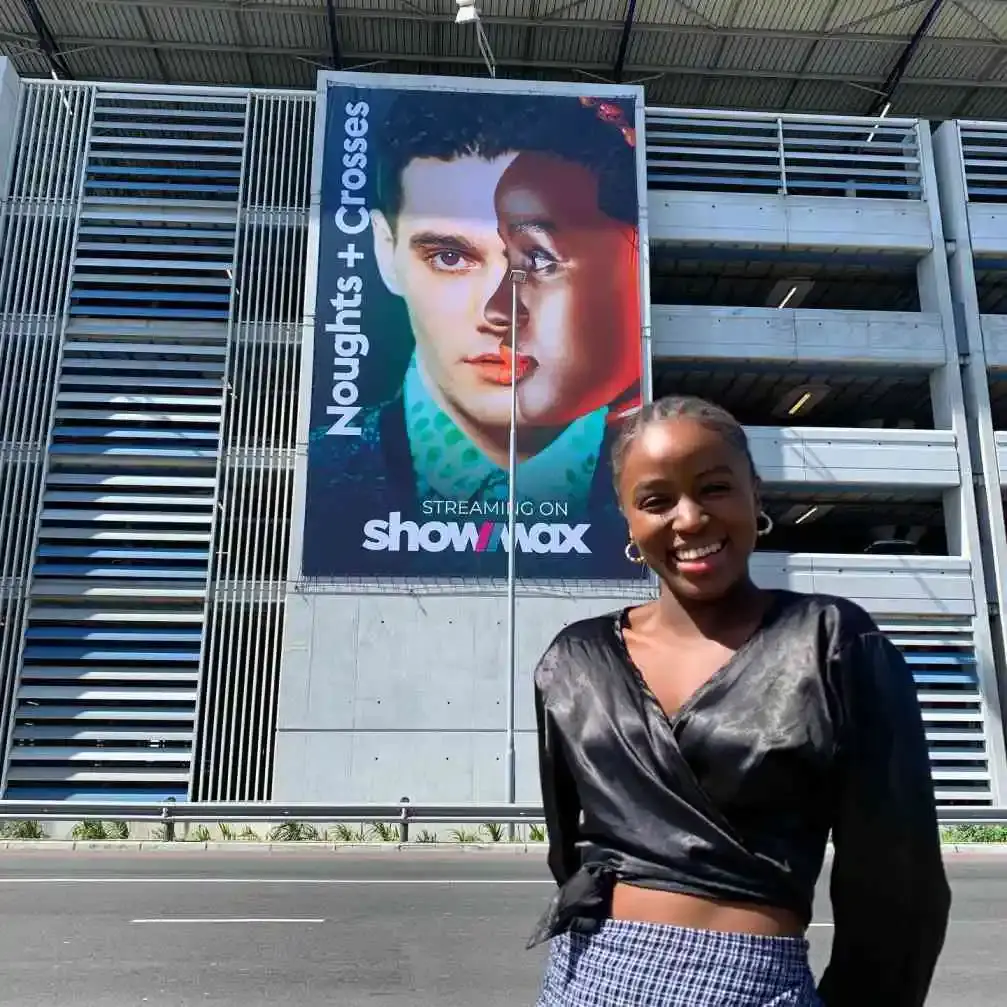 Masali Baduza smiles in front of a large "Noughts + Crosses" billboard featuring her face and co-star Jack Rowan.