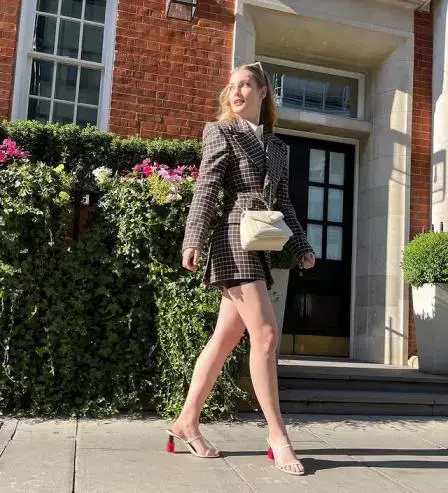 Hannah Dodd in a plaid blazer and heels walks past a brick building with greenery.