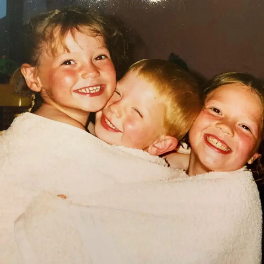 Hannah Dodd as a child smiling brightly while huddled together with her young siblings, wrapped in fluffy white towels.