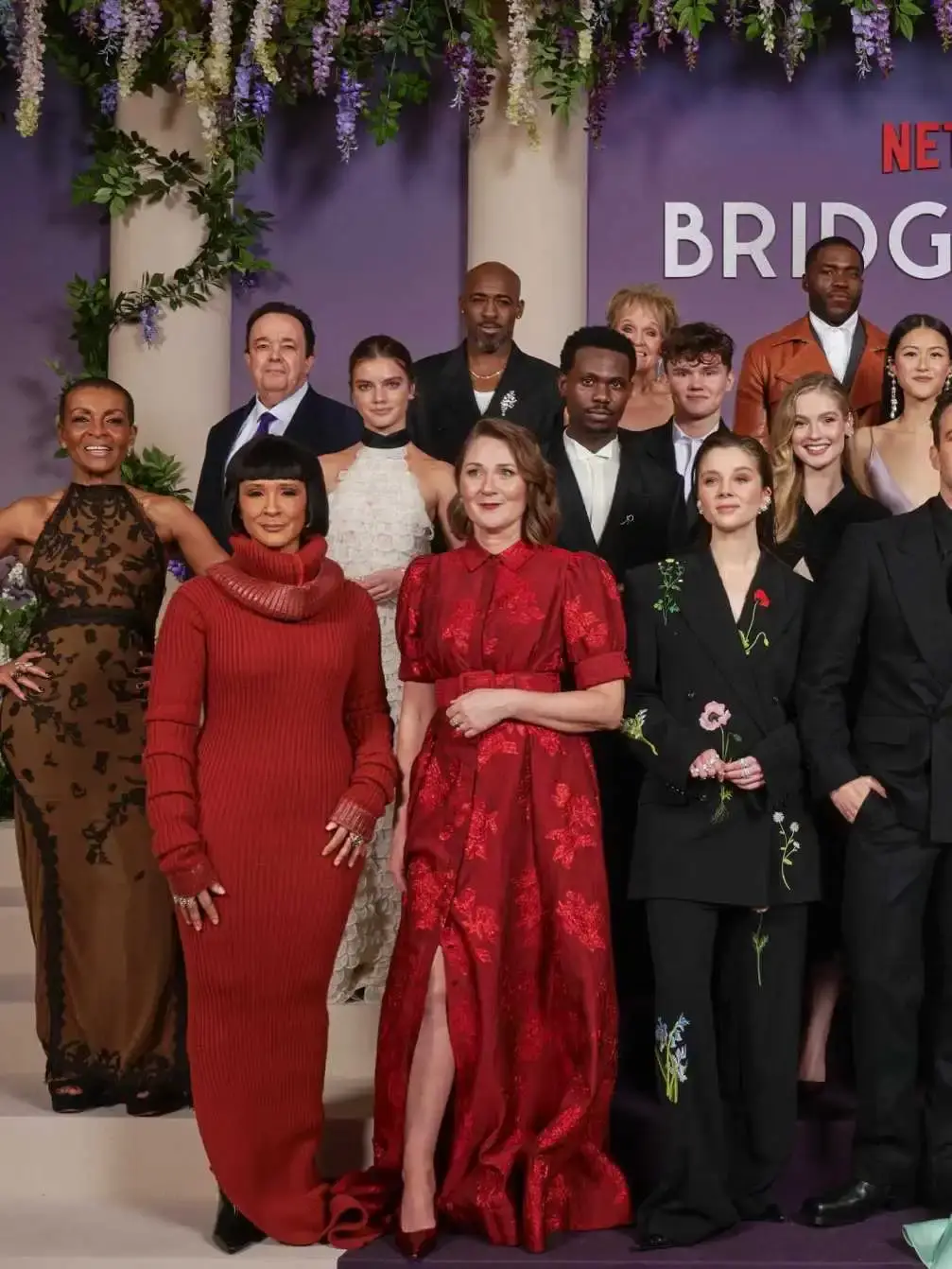 Victor Alli stands prominently among the ensemble cast of Bridgerton at a formal premiere event featuring a purple floral backdrop.