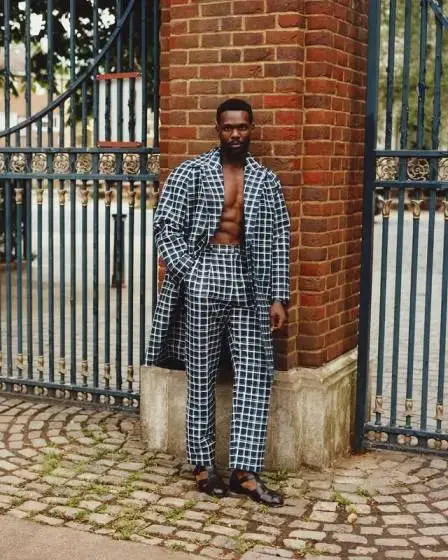 Victor Alli poses confidently by a brick pillar and iron gate wearing a stylish navy and white checkered suit with an open jacket.