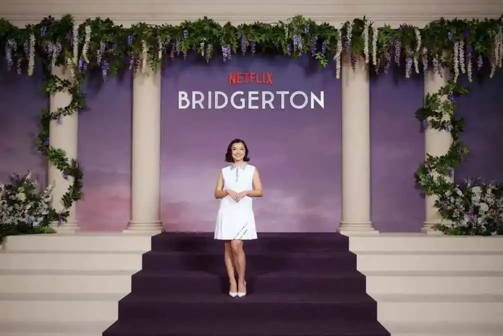 Michelle Mao smiles while posing in a white sleeveless dress on a purple stage in front of a Netflix Bridgerton backdrop decorated with floral greenery.