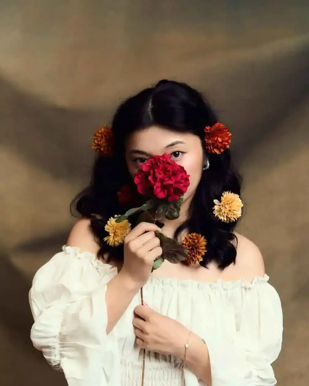 Michelle Mao holds a large red flower over her face while wearing an off-the-shoulder white top and yellow flowers in her hair against a brown studio backdrop.