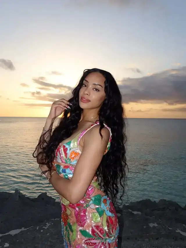 Content creator Kristy Scott poses in a colorful floral sequined dress against a scenic ocean sunset backdrop.
