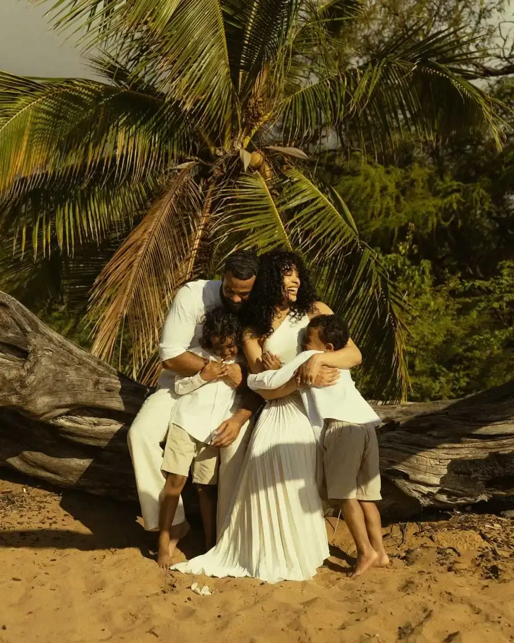 Kristy Scott and Desmond Scott share a joyful embrace with their two young sons while posing on a sandy beach in front of a large palm tree.