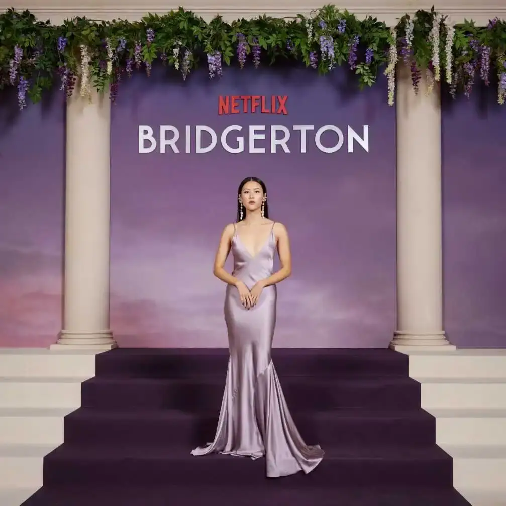 Isabella Wei stands on a purple carpeted staircase in a shimmering lilac slip gown in front of a Netflix Bridgerton backdrop.