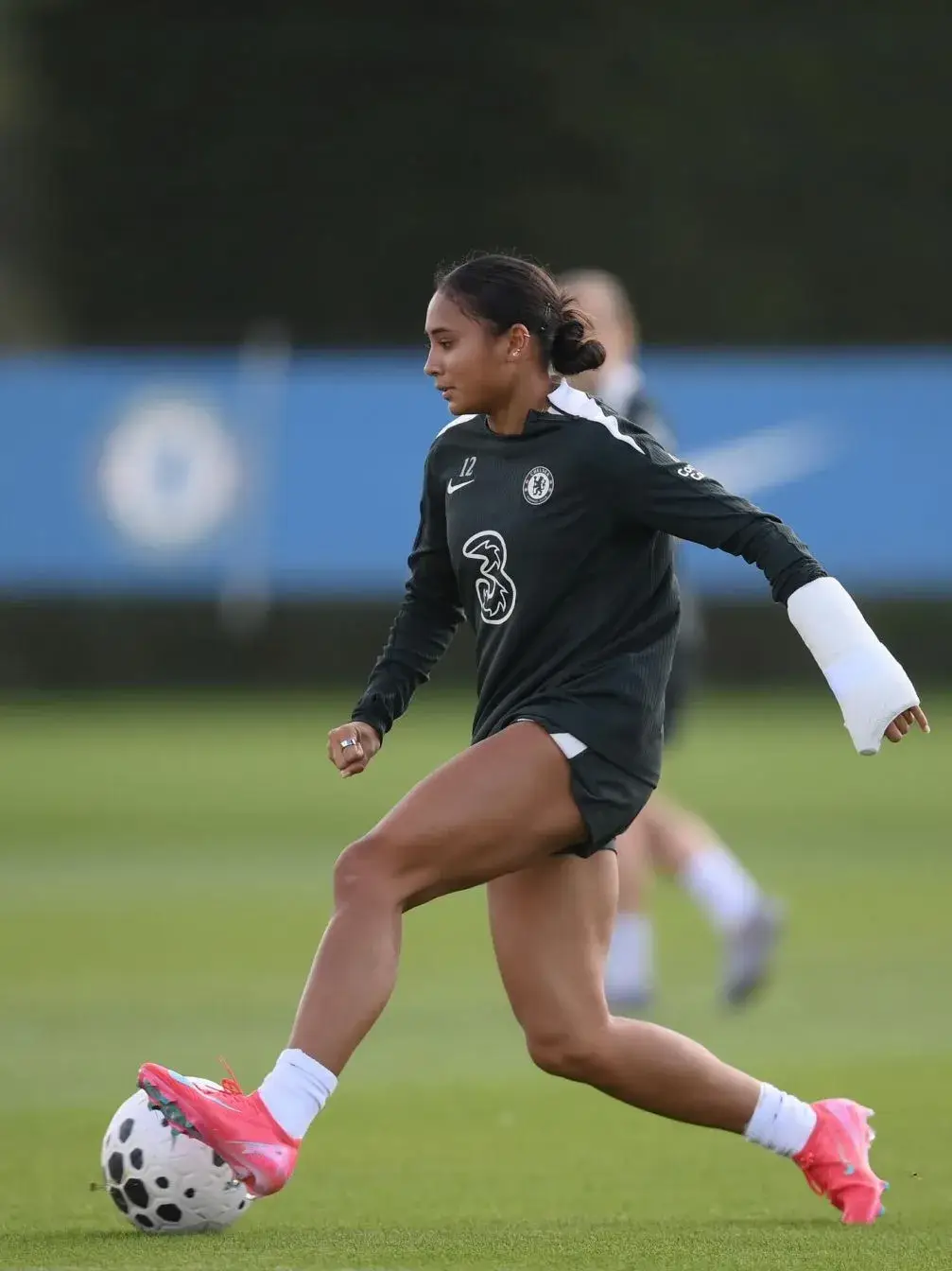 Soccer player Alyssa Thompson dribbles a ball across a green field while wearing a black Chelsea FC training kit and a protective white cast on her left arm.