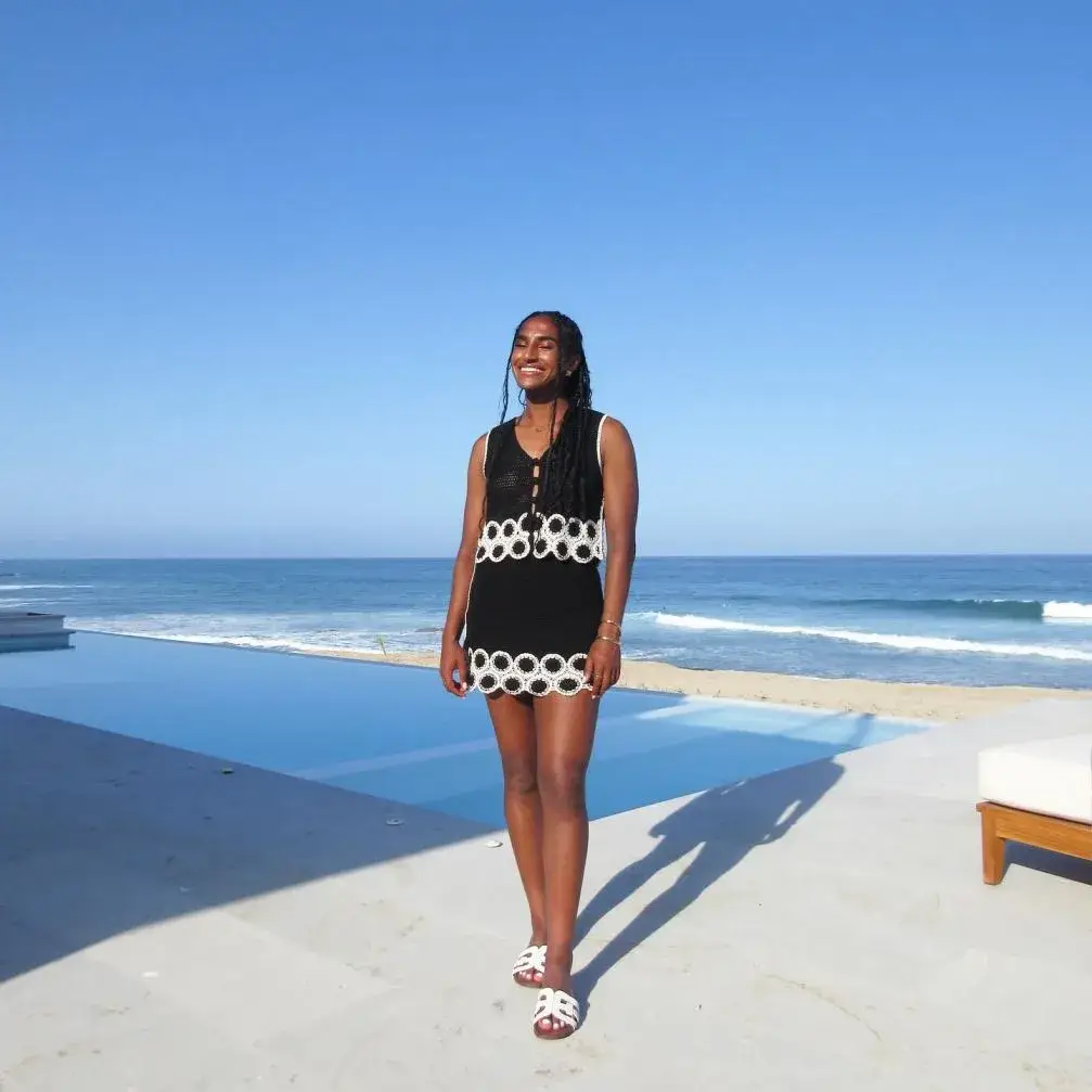 Naomi Girma stands smiling by a pool with the ocean in the background, wearing a black and white crochet dress.