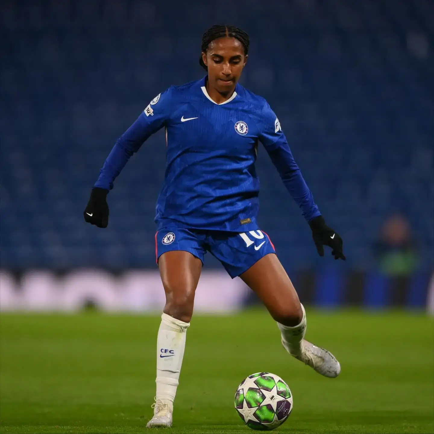 Naomi Girma dribbles a soccer ball across a grass field while wearing a blue Chelsea FC kit, white socks, and black gloves during a match.