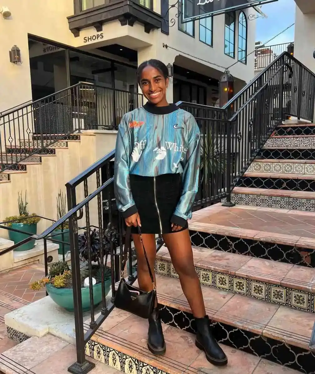 Naomi Girma stands on an outdoor tiled staircase with a joyful smile, wearing a metallic blue Off-White long-sleeve top, a black mini skirt, and black boots.