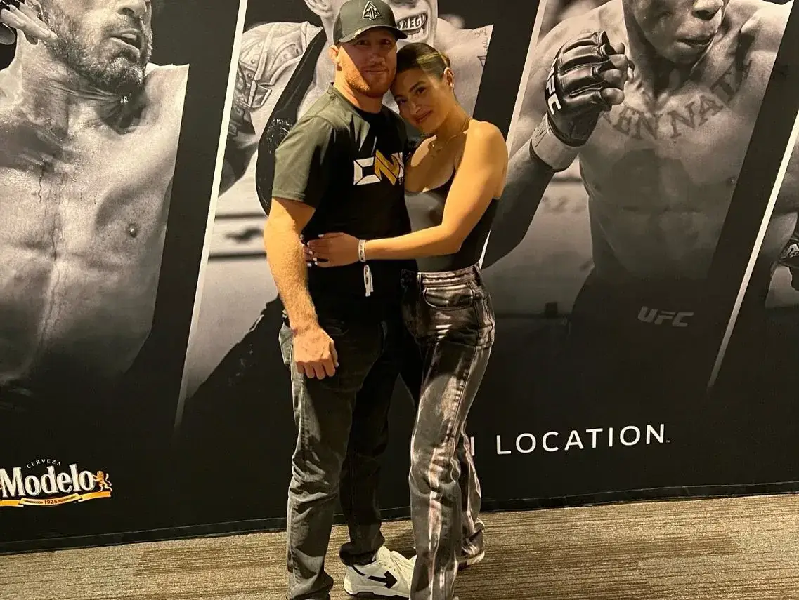 Justin Gaethje and his girlfriend, Sophia Romano, pose closely together in front of a UFC backdrop while Romano leans her head on his shoulder.
