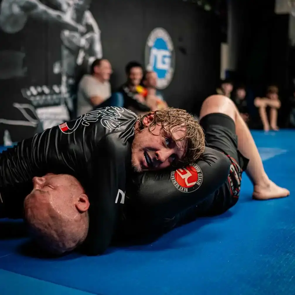 Paddy Pimblett smiles as he maintains a grappling hold on an opponent during a Jiu-Jitsu training session on a blue mat.
