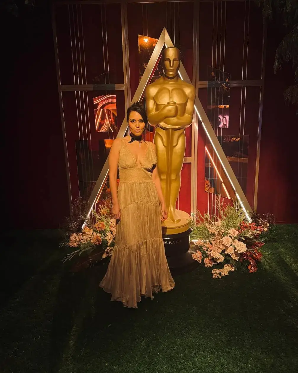 Actress Alyssa Diaz poses in a tiered gold gown next to a large Oscar statue at a formal event.