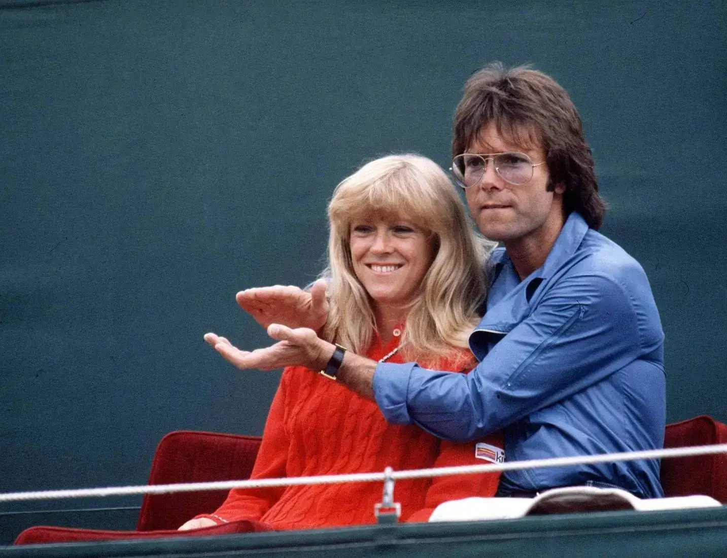 Pop star Cliff Richard and former tennis champion Sue Barker sit closely together in the stands at a tennis tournament in the early 1980s.