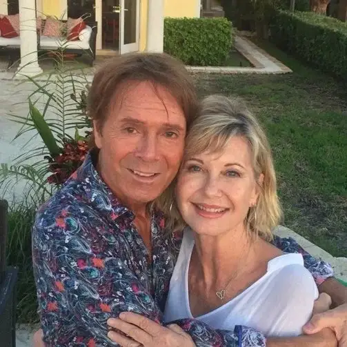 Sir Cliff Richard and Olivia Newton-John smile warmly while embracing for a photo outdoors in a garden setting.