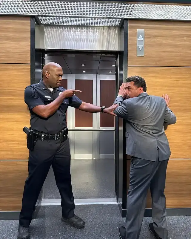 Actor Richard T. Jones, dressed in a dark police uniform, playfully points a finger at a man in a grey suit while standing in front of an elevator.