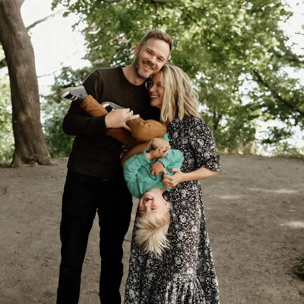 Actor Shawn Ashmore and his wife Dana Renee smile and laugh while holding their young son, Oliver, upside down during a family outing in a wooded park.