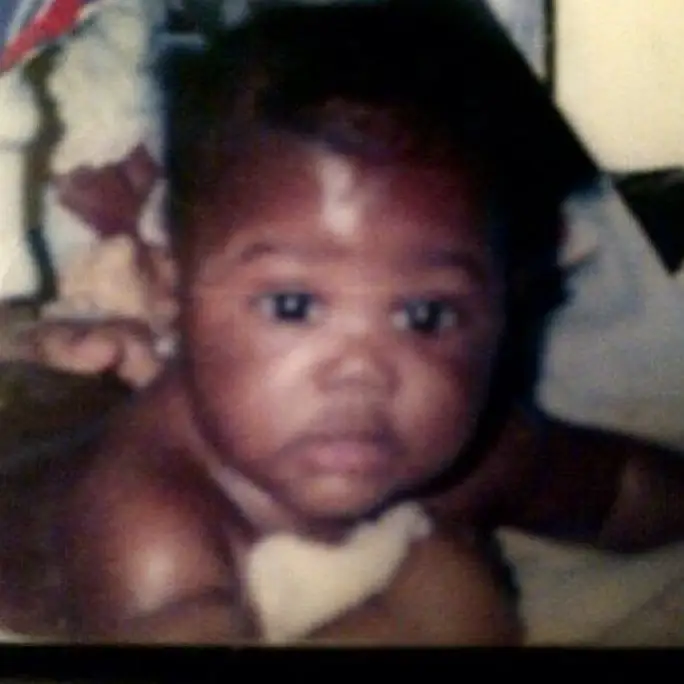 A close-up, slightly blurry childhood photograph shows a young Afton Williamson as a baby looking directly at the camera while resting on a soft surface.