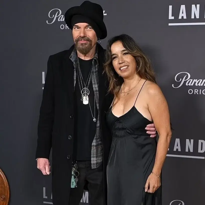 Actor Billy Bob Thornton, in a black coat and hat with silver necklaces, stands with his arm around Connie Angland, who is wearing a black spaghetti-strap dress, on a red carpet.
