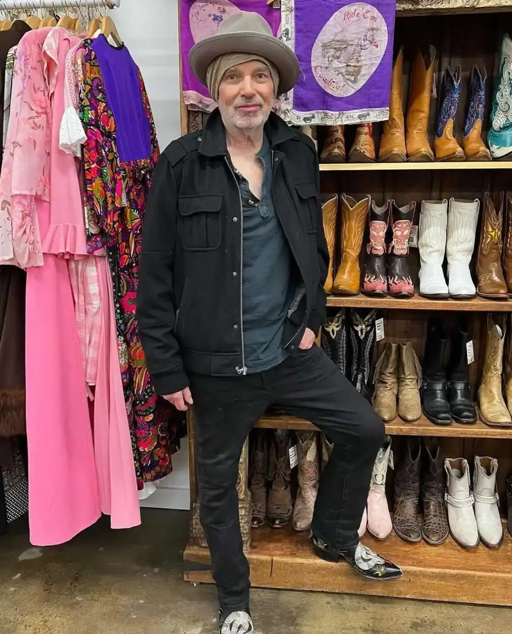 Actor Billy Bob Thornton stands in a vintage clothing shop wearing a black jacket and a grey fedora while leaning against a shelf filled with cowboy boots.