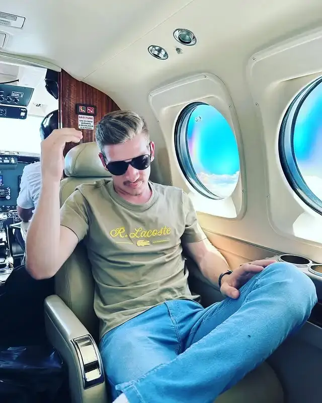 Actor Gabriel Basso sits in a tan leather seat inside a private airplane while wearing a green t-shirt, blue jeans, and dark sunglasses.