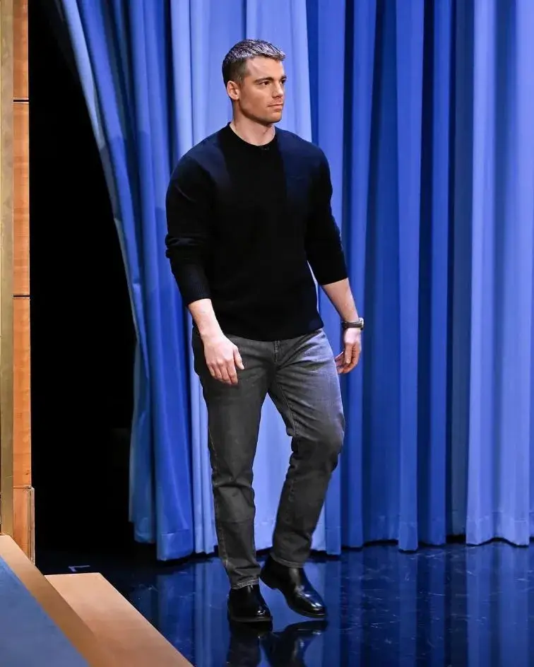 Actor Gabriel Basso walks across a stage in a black long-sleeved shirt and grey jeans against a backdrop of blue curtains.