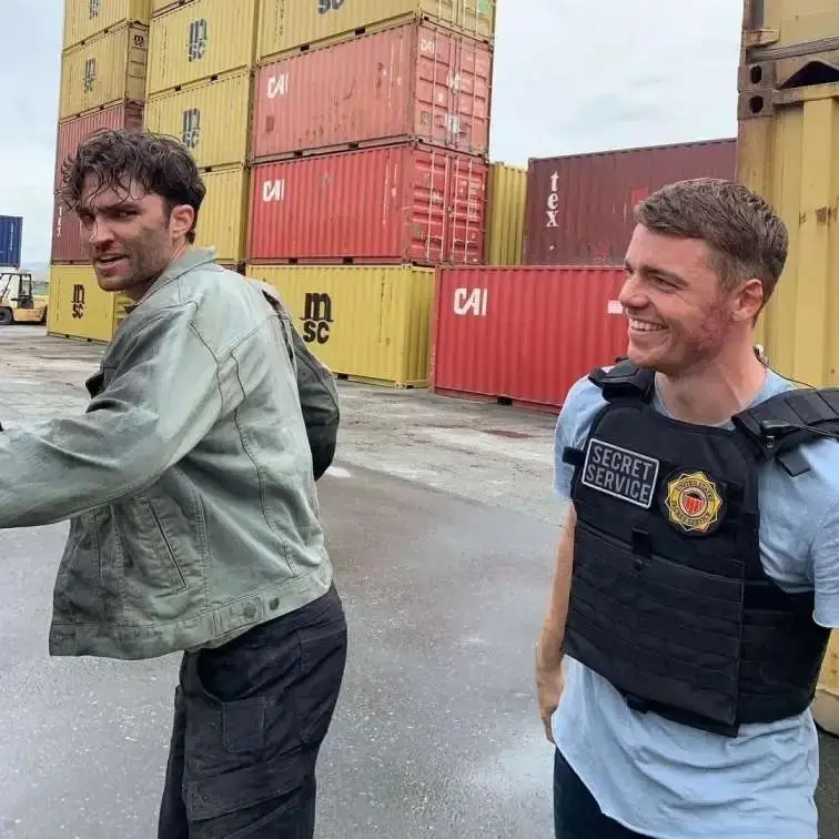 Actor Gabriel Basso and co-star stand together in front of stacked shipping containers on the outdoor set of The Night Agent.