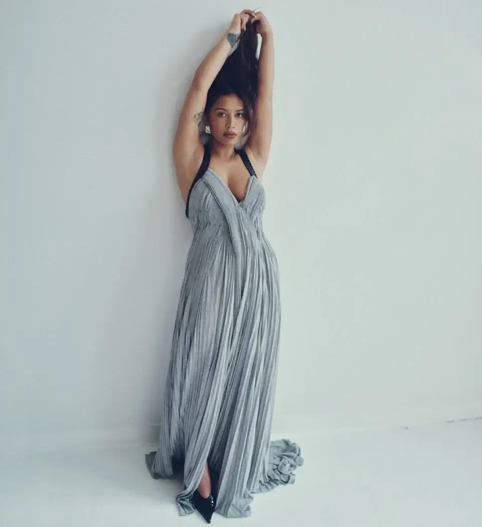Actor Luciane Buchanan poses against a plain white wall while wearing a floor-length, pleated grey gown with black straps.