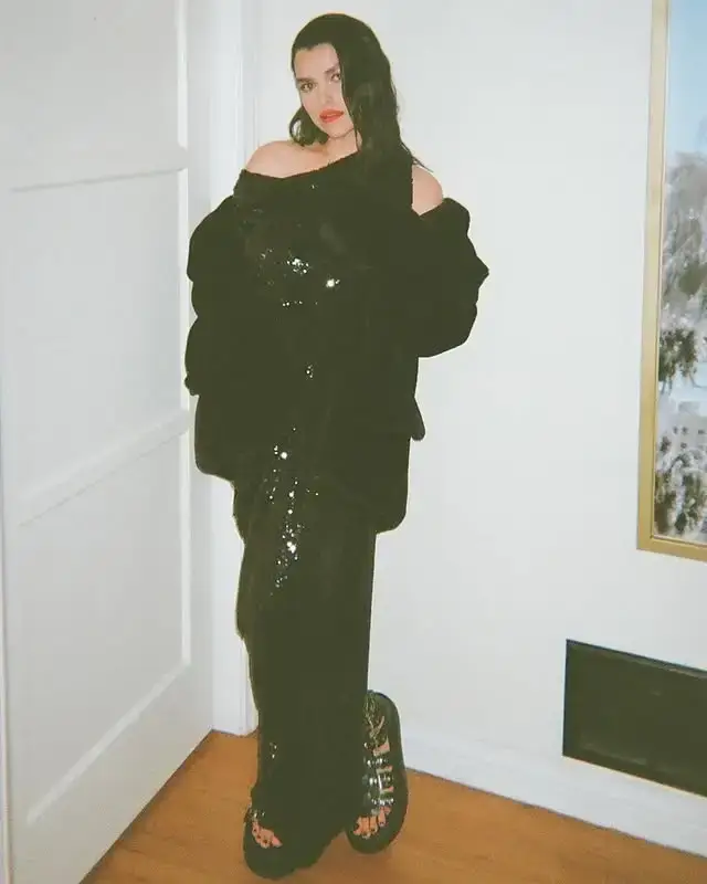 Actor Eve Harlow poses in a full-length photo wearing a shimmering black sequined gown and platform sandals against a plain white wall.