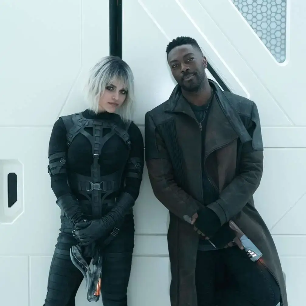 Alt Text: Actors Eve Harlow and David Ajala, portraying the characters Moll and Cleveland "Book" Booker, stand together on the futuristic set of Star Trek: Discovery.