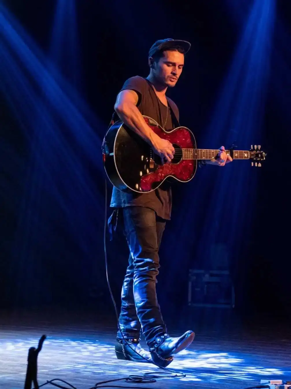 Actor and musician Michael Malarkey, known for his role as Enzo in The Vampire Diaries, poses thoughtfully in a dark denim jacket with his hands clasped.