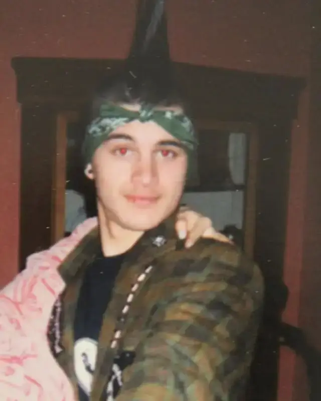 Young Michael Malarkey with a tall, dark mohawk and a green patterned bandana posing in a camouflage jacket.