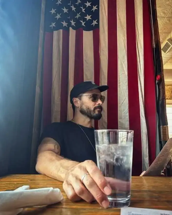 Michael Malarkey sits at a wooden table holding a glass of water with a large American flag hanging in the background.