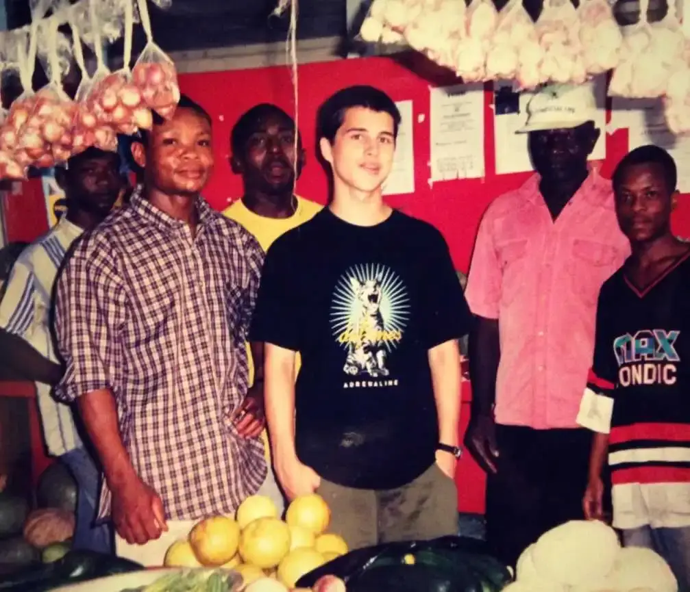 A young Michael Malarkey stands in a black graphic t-shirt among a group of men at an outdoor market stall with hanging produce.