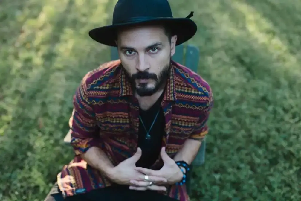 Michael Malarkey sits outdoors in a patterned shirt and a black wide-brimmed hat with his hands clasped together.