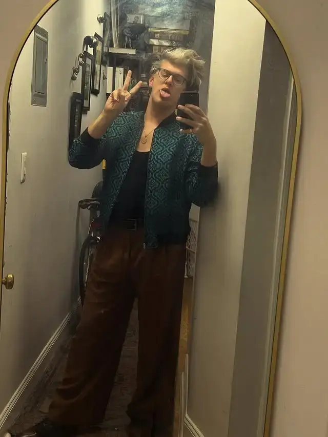 James Ortiz takes a mirror selfie in a hallway while wearing a teal patterned jacket, brown trousers, and making a peace sign.